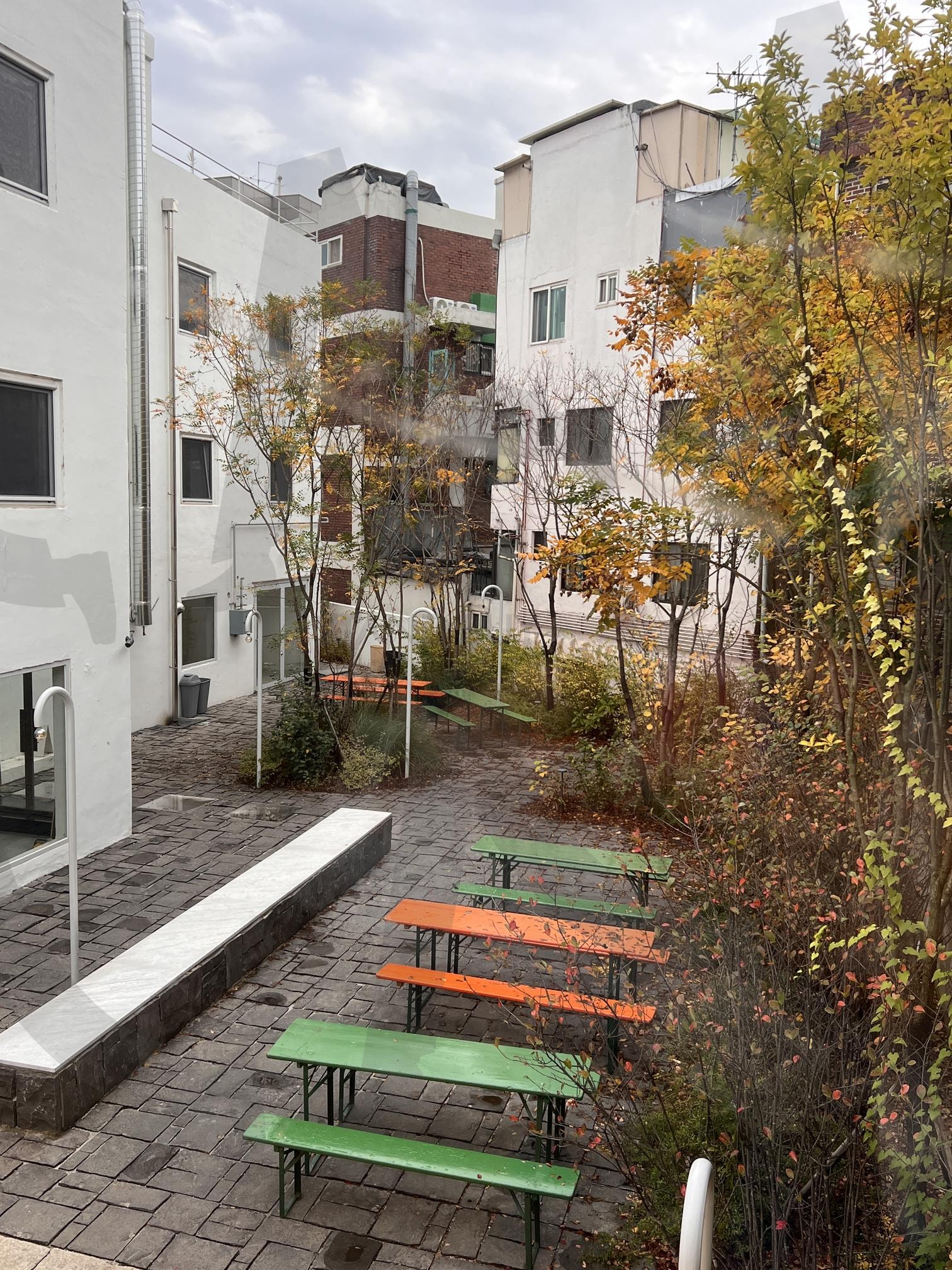 젖은 돌 바닥의 야외 테이블과 벤치 | 야외 테이블, 벤치, 돌 바닥, 젖은, 비, 흐린 날씨, 나무, 건물, 도시, 가을 | 스탠바이 STANDBUY 디지털 콘텐츠